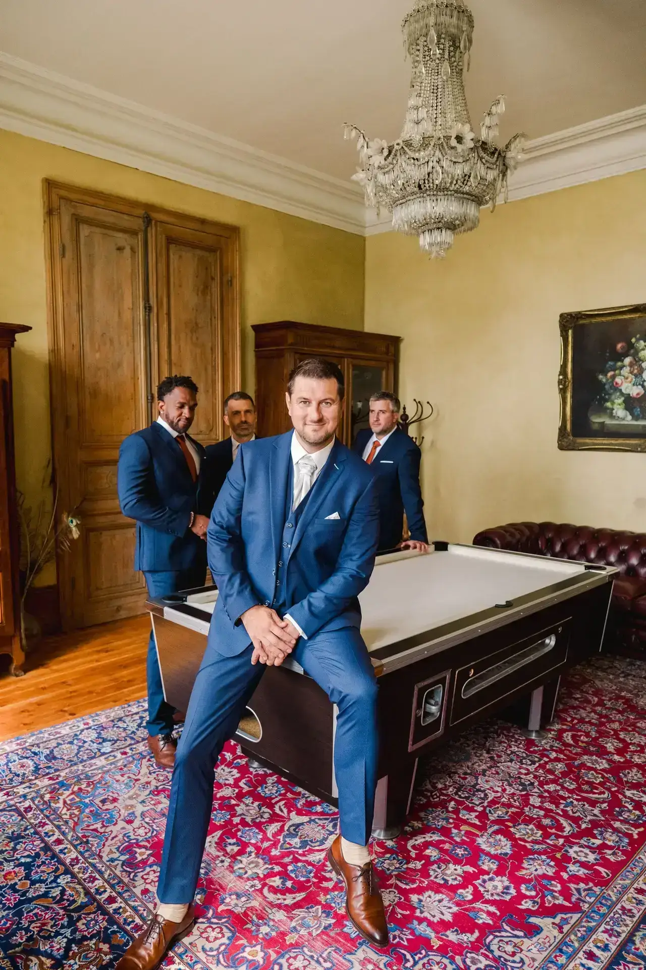 Portrait élégant du marié et de ses témoins, réunis autour d’une table de billard sous un lustre ancien, dans l’atmosphère feutrée du château.