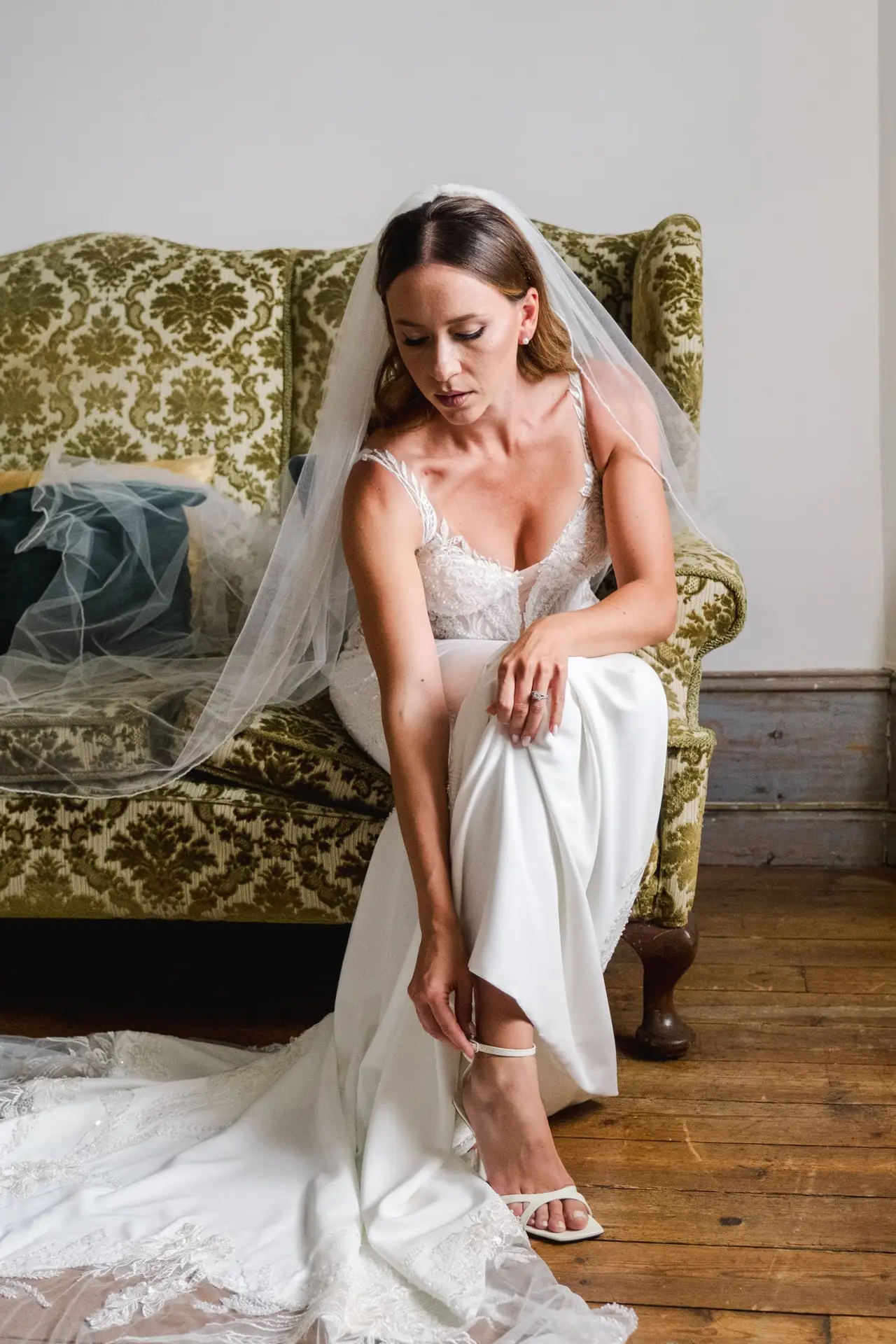 Mariée en train de se préparer au Château Fengari près de Bordeaux, assise sur un fauteuil ancien, ajustant sa sandale, portant une robe de mariée élégante en dentelle.