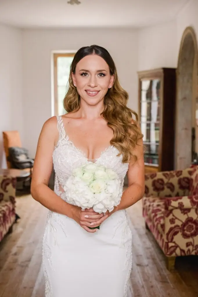 Portrait de la mariée debout à l’intérieur d’un salon élégant et lumineux, regardant directement l’objectif avec un sourire doux et confiant. Elle porte une robe de mariée blanche finement travaillée avec de la dentelle florale et des broderies délicates, mettant en valeur le haut du buste. Ses cheveux sont coiffés en ondulations souples et naturelles, tombant sur ses épaules, et elle tient un bouquet rond de fleurs blanches, composé d’hortensias et de roses, entre ses deux mains. En arrière-plan, on distingue des canapés anciens aux motifs floraux, un sol en bois clair et une bibliothèque vitrée, créant une ambiance chaleureuse et raffinée. L’image évoque l’élégance, la sérénité et l’émotion d’un moment clé de la journée de mariage.