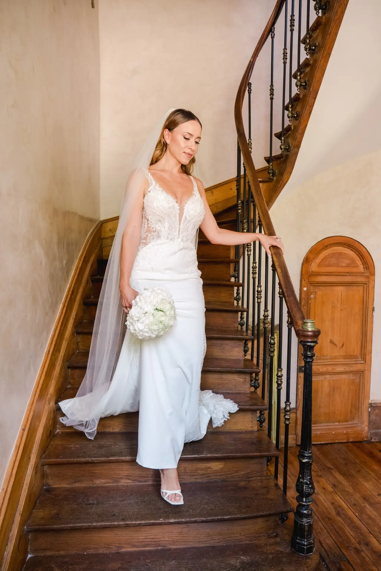 Mariée descendant lentement un élégant escalier en bois ancien à l’intérieur d’un château ou d’une demeure historique, vêtue d’une robe de mariée blanche ajustée avec un bustier en dentelle florale et un décolleté en V, portant un long voile délicat qui tombe le long de l’escalier. Elle tient un bouquet rond composé de fleurs blanches, probablement des hortensias et des roses, dans sa main gauche, tandis que sa main droite repose sur la rampe en bois sculpté et métal. L’escalier en colimaçon est éclairé par une lumière douce et naturelle, mettant en valeur les murs patinés et le parquet ancien. L’atmosphère est élégante, intime et chargée d’émotion, illustrant un moment calme et solennel avant la cérémonie de mariage.