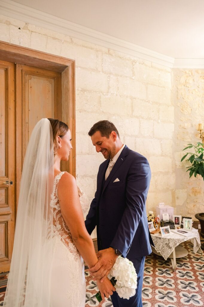 Moment intime entre les mariés à l’intérieur du Château Fengari, où ils se tiennent face à face en se tenant les mains. Le marié sourit avec émotion tandis que la mariée, en robe de dentelle et voile long, lui fait face. À l’arrière-plan, une table décorée avec des photos et objets personnels rappelle l’importance des souvenirs et de la famille lors de cette journée de mariage.