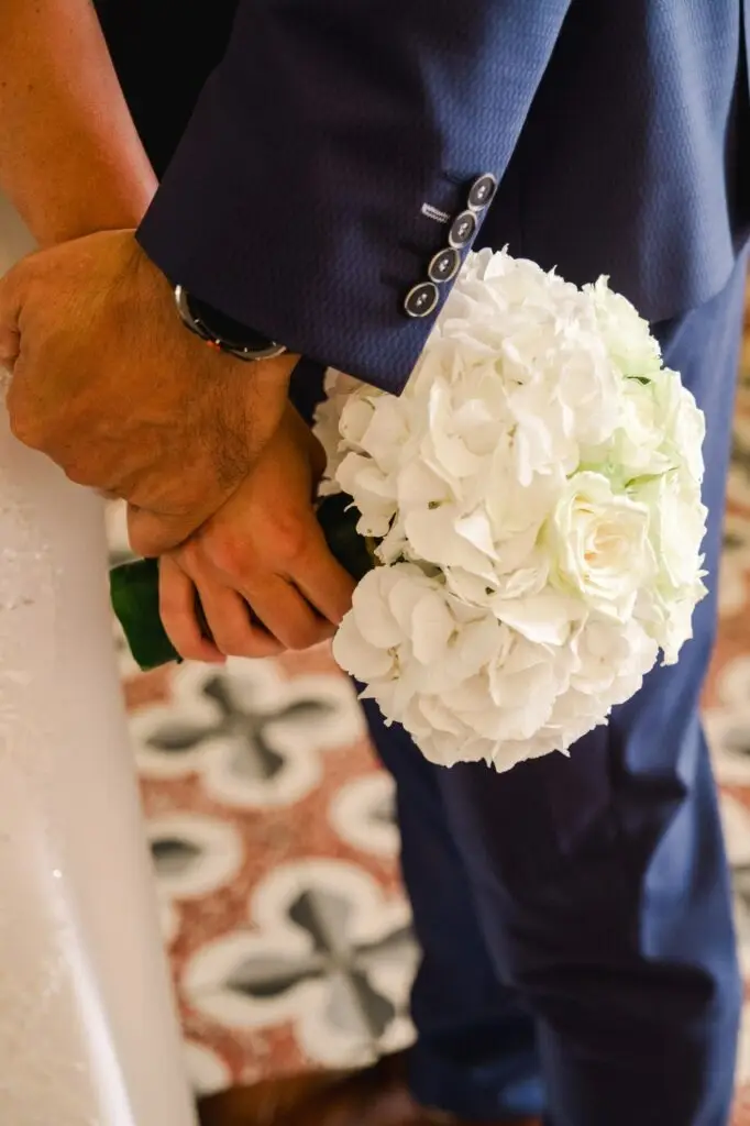 Gros plan sur les mains des mariés tenant ensemble un bouquet de fleurs blanches, composé principalement d’hortensias et de roses. Le costume bleu du marié contraste avec la robe claire de la mariée, tandis que les textures du tissu et des fleurs apportent une sensation d’intimité et de tendresse, capturant un moment symbolique du mariage au Château Fengari.