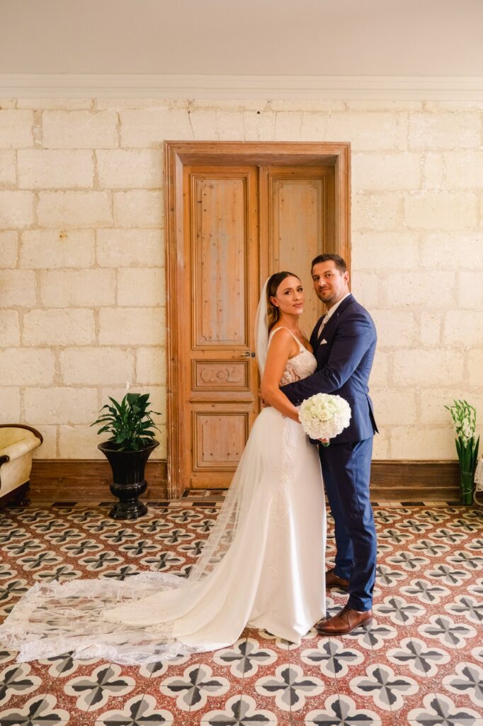 Portrait des mariés posant ensemble dans un couloir du Château Fengari à Bordeaux, devant une grande porte en bois ancien. La mariée porte une robe de mariage en dentelle avec une longue traîne et un voile délicat, tandis que le marié est vêtu d’un costume bleu élégant et tient un bouquet de fleurs blanches. L’ambiance est douce et intemporelle, mettant en valeur l’architecture en pierre du château et l’élégance du couple.