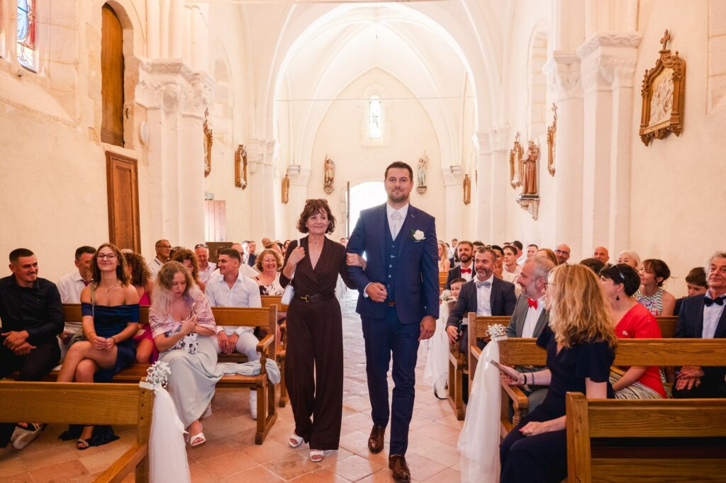 Le marié avance dans l’allée centrale de l’église accompagné d’une femme, probablement un membre de sa famille, qui le tient par le bras. Ils marchent entre les bancs occupés par les invités, qui les regardent avec attention et sourires. Le marié porte un costume bleu élégant, tandis que sa compagne est vêtue d’une tenue sombre. L’église est lumineuse, avec une architecture en pierre, des statues religieuses le long des murs et une atmosphère chaleureuse et cérémoniale.