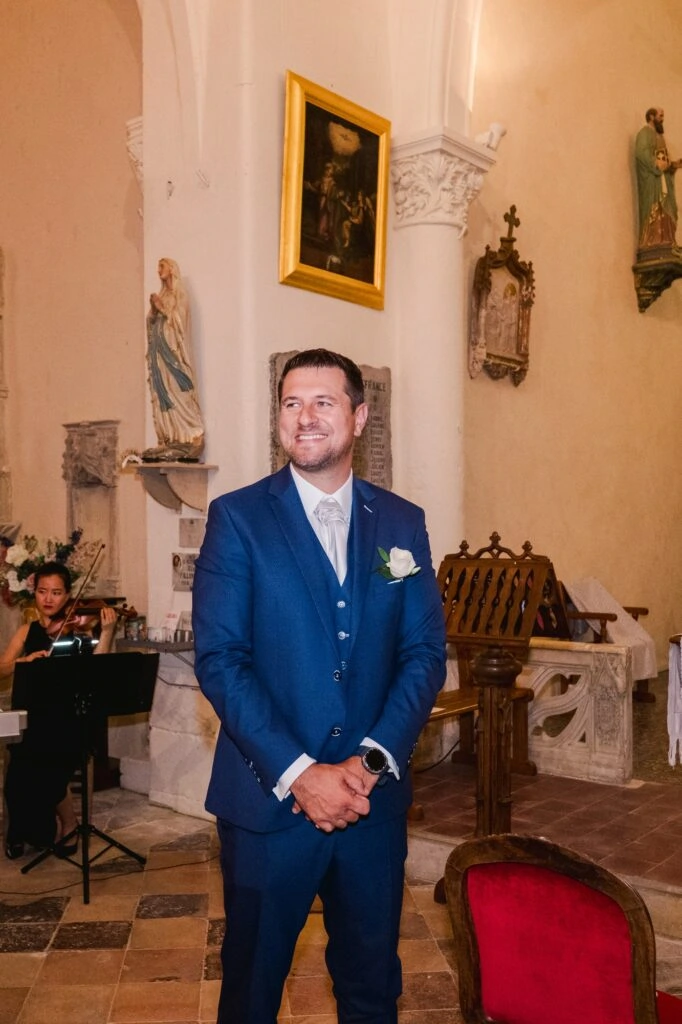 Le marié se tient debout près de l’autel à l’intérieur de l’église, les mains jointes devant lui, souriant avec émotion. Il porte un costume bleu trois-pièces avec une cravate claire, une boutonnière blanche et une montre au poignet. En arrière-plan, on distingue des éléments religieux, un pupitre en bois sculpté, des statues et une musicienne jouant du violon, contribuant à l’ambiance solennelle et élégante de la cérémonie de mariage.