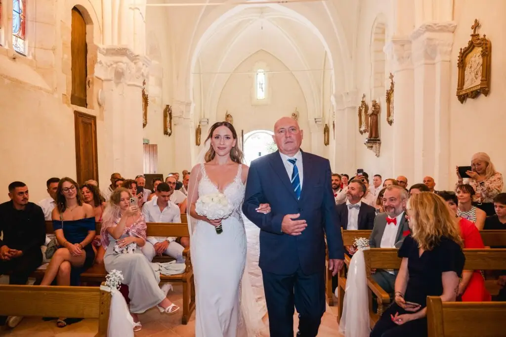 La mariée avance lentement dans l’allée centrale de l’église, tenant son bouquet de fleurs blanches, le bras enlacé avec un homme plus âgé portant un costume sombre et une cravate bleue. Les invités sont assis de chaque côté sur des bancs en bois, certains souriant, d’autres prenant des photos avec leur téléphone. L’architecture de l’église est mise en valeur par une voûte haute, des colonnes en pierre claire et des statues religieuses accrochées aux murs, créant une atmosphère solennelle et émouvante.