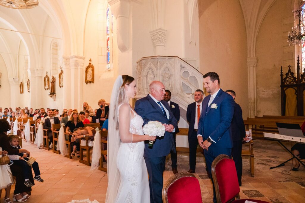 a mariée, vêtue d’une robe de mariée blanche ajustée avec un bustier en dentelle et un long voile transparent, se tient dans l’allée centrale de l’église aux côtés d’un homme plus âgé en costume bleu foncé, probablement son père. Elle tient un bouquet rond de fleurs blanches. Face à eux, le marié, habillé d’un costume bleu élégant avec une cravate claire et une boutonnière blanche, les regarde avec émotion. L’intérieur de l’église est lumineux, avec des murs clairs en pierre, des statues religieuses fixées aux colonnes et des invités assis sur des bancs en bois décorés de tissus blancs.