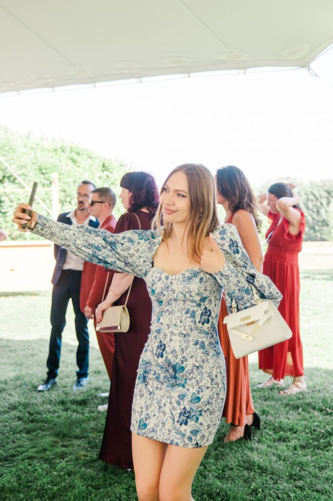 Une invitée élégamment habillée prend un selfie avec son téléphone pendant le cocktail d’un mariage au Château Fengari, près de Bordeaux. Elle se tient sous la tente de réception, entourée d’autres invités en arrière-plan, dans une ambiance festive et détendue. Cette photo de mariage capture un moment spontané et moderne de la réception, illustrant la joie et la convivialité de la journée, photographiée par un photographe de mariage à Bordeaux.