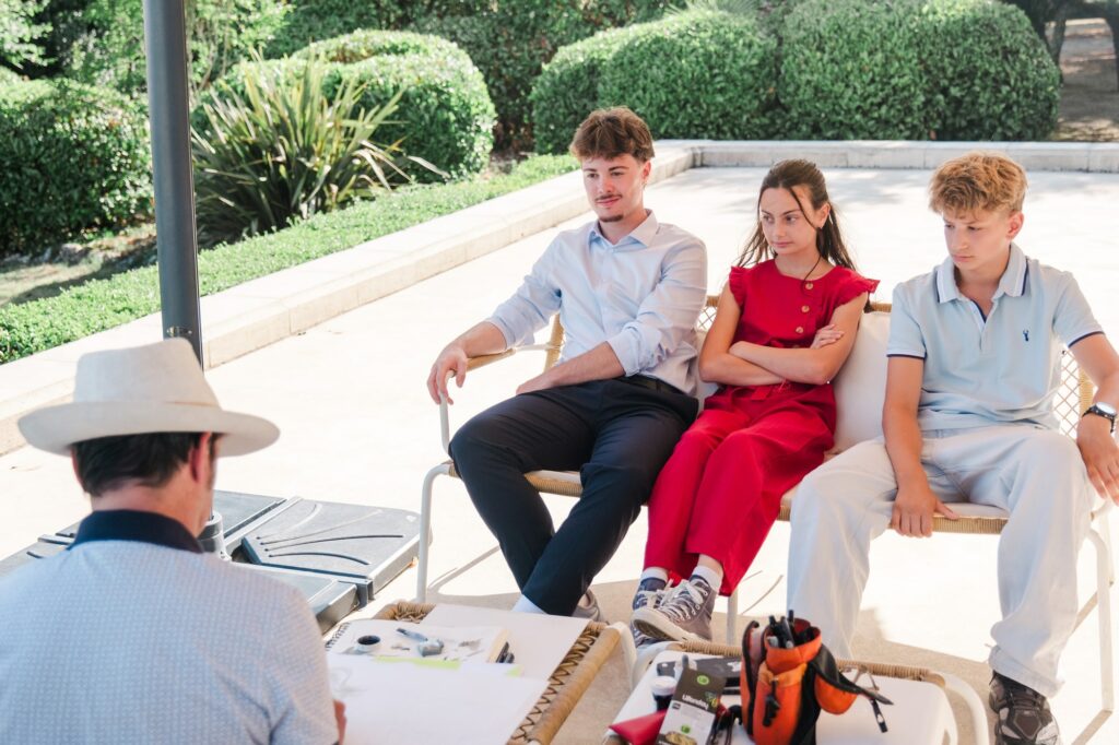Trois jeunes invités assis côte à côte posent calmement pendant qu’un caricaturiste réalise leurs portraits lors d’un mariage au Château Fengari, près de Bordeaux. L’animation se déroule en extérieur, dans le parc du château, sous une lumière naturelle douce. Cette photographie de mariage met en avant les moments de détente et de partage vécus par les invités, capturés par un photographe de mariage en Gironde attentif aux scènes de vie.