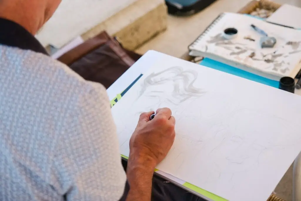 Un artiste caricaturiste en train de dessiner pendant le cocktail d’un mariage au Château Fengari, près de Bordeaux. La photo montre la main de l’artiste esquissant les visages des invités sur une feuille de papier, avec le matériel de dessin posé sur la table. Cette scène illustre une animation originale et conviviale, apportant une touche artistique au mariage. Une image prise par un photographe de mariage à Bordeaux, capturant les moments spontanés et créatifs de la réception.
