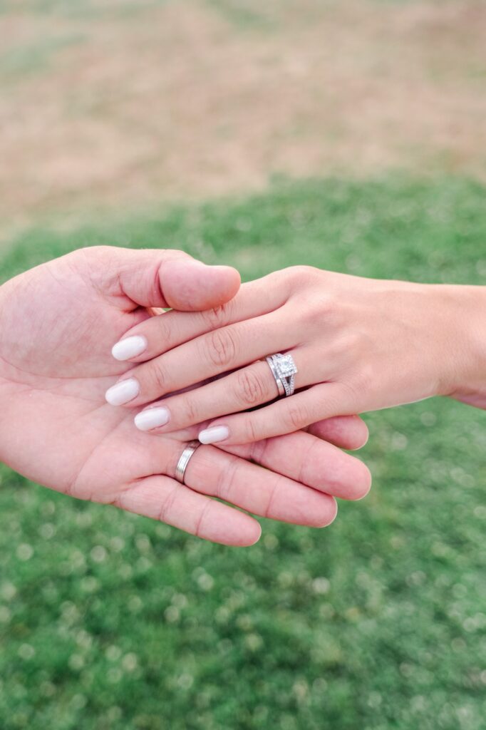 Gros plan sur les mains des mariés se touchant délicatement, mettant en évidence leurs alliances. La main de la mariée porte une bague sertie d’un diamant brillant, accompagnée d’une alliance assortie, tandis que la main du marié arbore une alliance simple et élégante. Les ongles de la mariée sont soignés, d’une teinte claire et naturelle. L’arrière-plan flou de verdure met en valeur les bijoux, symboles forts de l’engagement et de l’union du couple.
