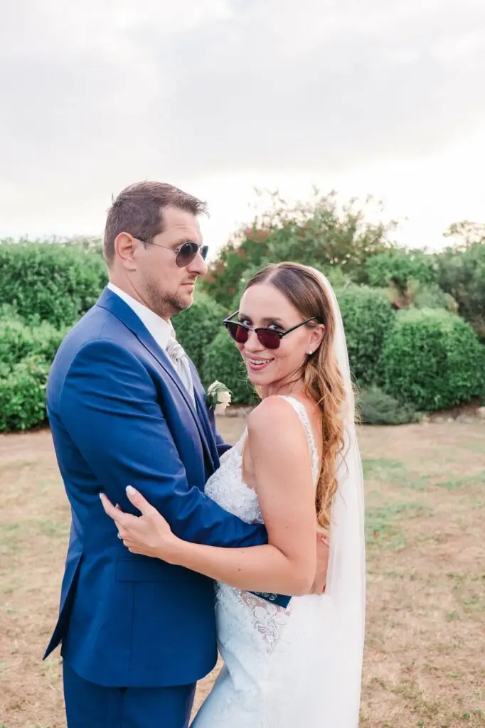 Le couple de mariés pose de manière décontractée dans un jardin, tous deux portant des lunettes de soleil. Le marié, en costume bleu, adopte une expression légèrement sérieuse et amusée, tandis que la mariée sourit en regardant l’objectif par-dessus ses lunettes. Sa robe de mariée blanche en dentelle et son voile apportent une touche de raffinement à cette scène plus légère et moderne. L’arrière-plan végétal et la lumière naturelle renforcent l’ambiance estivale et joyeuse, illustrant un moment spontané et plein de personnalité.