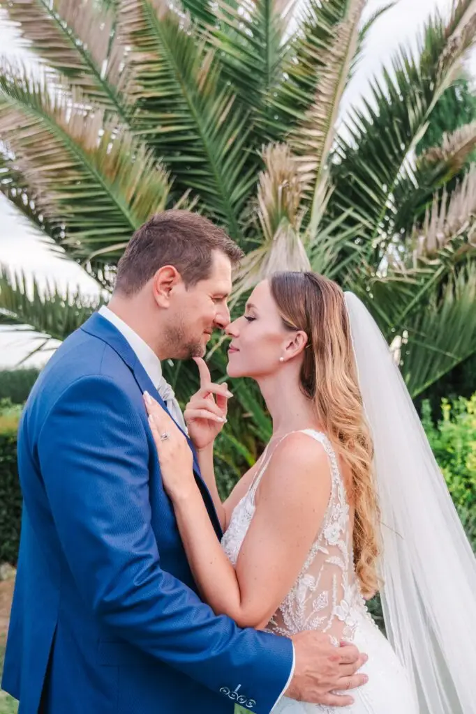 Les mariés se tiennent face à face dans un jardin, entourés de grands palmiers verdoyants. Le marié, en costume bleu, enlace la mariée qui porte une robe de mariée dos nu richement décorée de dentelle florale. La mariée pose délicatement un doigt sur le menton de son époux, dans un geste tendre et complice. Les visages sont très proches, presque sur le point de s’embrasser, créant une scène romantique et intime. Le décor naturel, baigné de lumière douce, accentue l’émotion et l’élégance de ce moment capturé lors d’une séance photo de mariage en extérieur.