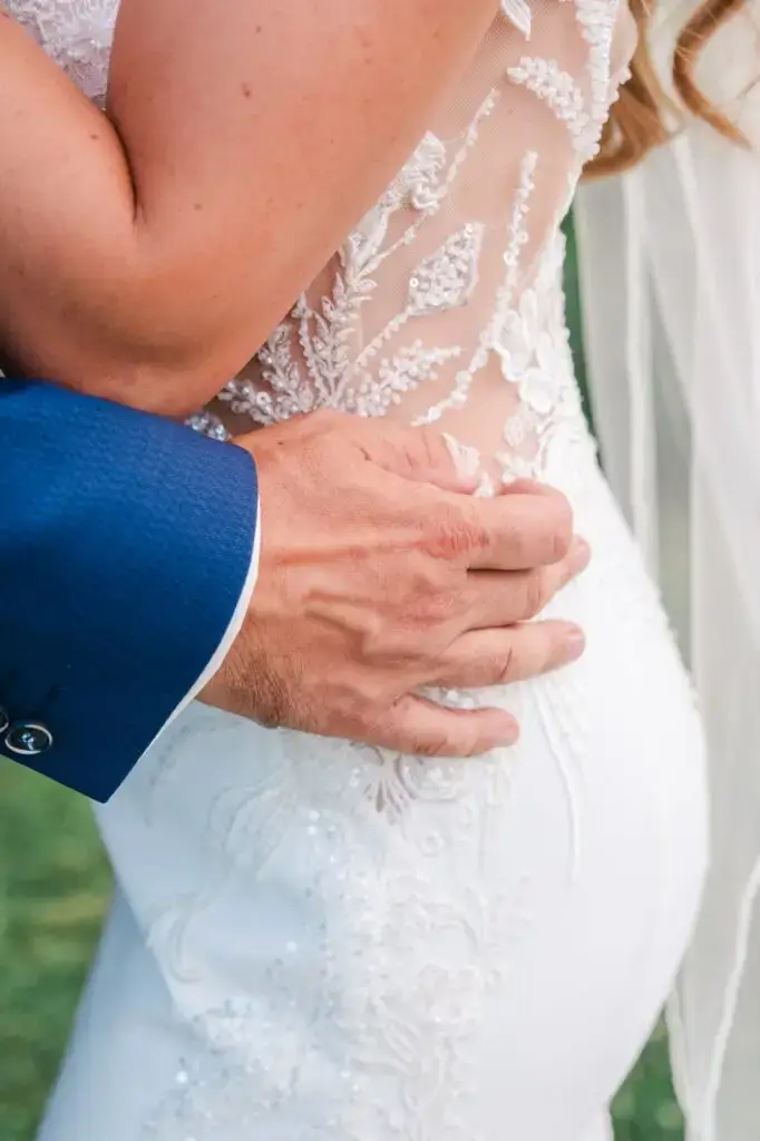 Gros plan détaillé montrant la main du marié posée avec tendresse sur la taille de la mariée. La manche du costume bleu contraste avec la robe blanche ornée de broderies florales et de perles délicates. La texture fine de la dentelle est clairement visible, soulignant le travail artisanal de la robe. Cette image intime met en avant la proximité du couple et les détails élégants des tenues, symbolisant la douceur et l’engagement du moment.