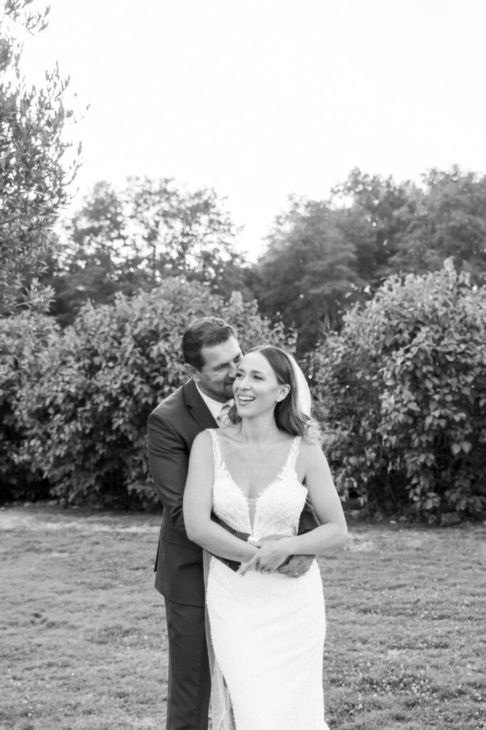 Photographie de mariage en noir et blanc montrant un couple enlacé dans un jardin. La mariée est au premier plan, souriante, portant sa robe de mariée en dentelle avec un décolleté délicat, tandis que le marié se tient derrière elle, les bras autour de sa taille, le visage proche du sien dans un geste tendre. Le décor extérieur est composé d’arbustes et d’arbres légèrement flous, ce qui concentre l’attention sur l’intimité et la complicité du couple. L’image transmet une forte émotion de bonheur et de proximité.