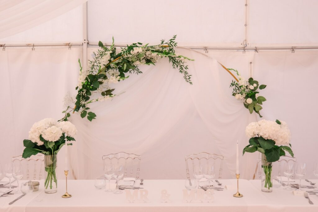 Vue de la table d’honneur décorée de fleurs blanches et de feuillage sous la tente de réception du Château Fengari, près de Bordeaux, mise en scène élégante du repas de mariage photographiée par un photographe de mariage.