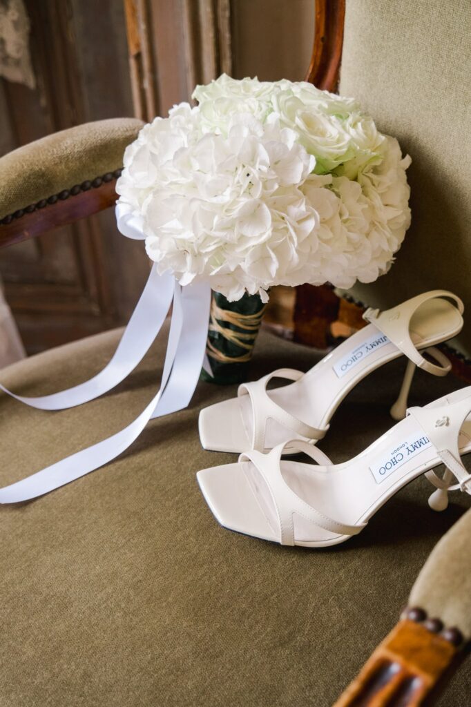 Détails raffinés des préparatifs de la mariée : bouquet de fleurs blanches et chaussures élégantes, posés sur un fauteuil ancien.