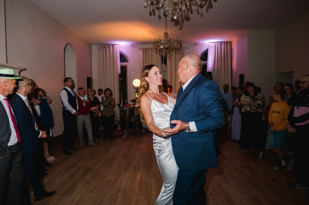 Photo d’un couple en train de danser lors d’un mariage, entouré d’invités, ambiance chaleureuse et lumineuse, chandeliers, décoration raffinée, moments précieux.