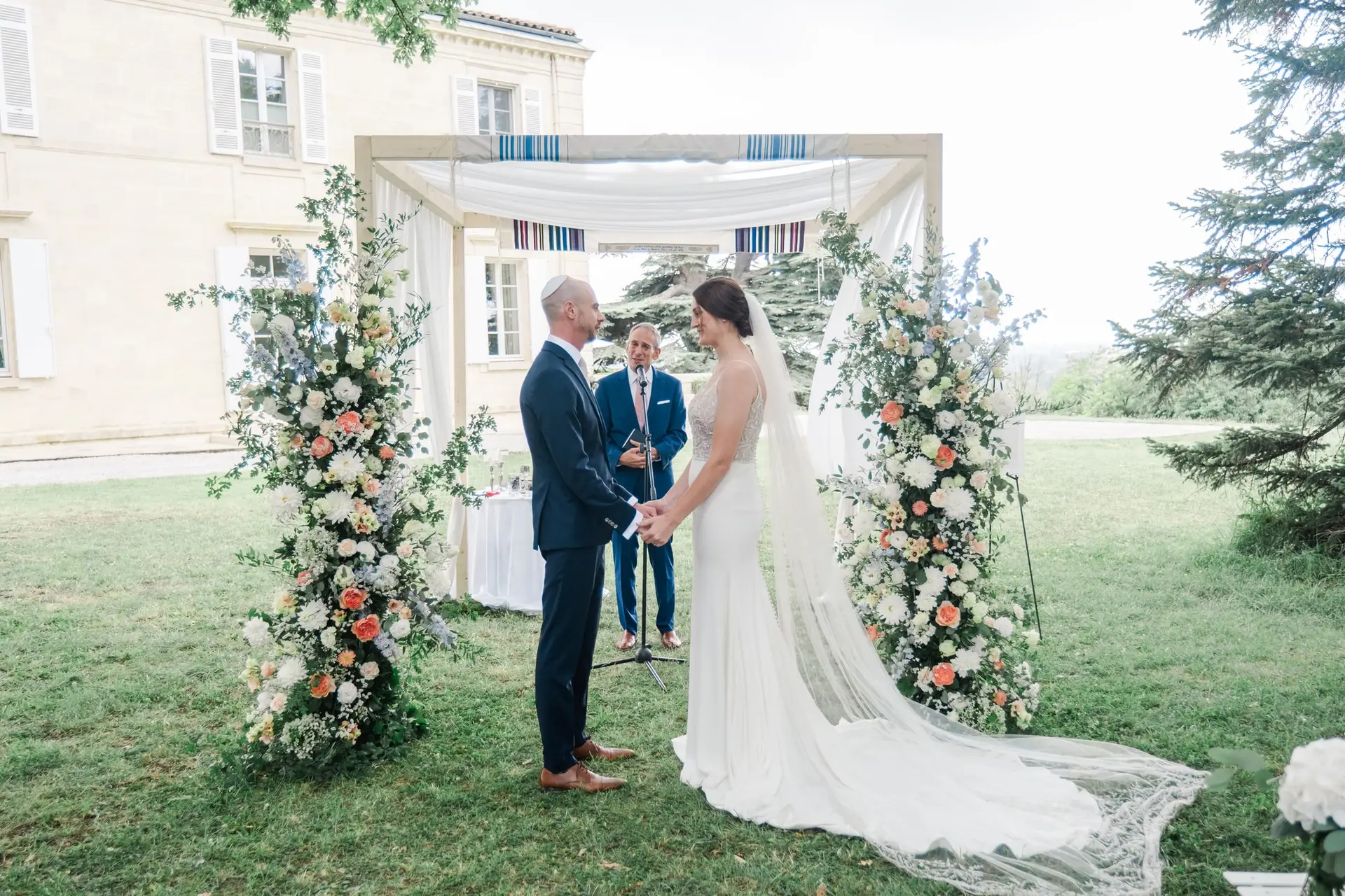 Les mariés se tenant la main sous l’arche fleurie pendant la cérémonie de mariage au Château Gassies à Bordeaux.