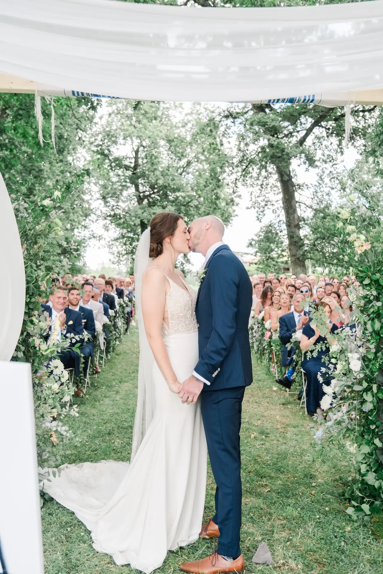 Les mariés s’embrassent sous une arche fleurie devant leurs invités lors de la cérémonie de mariage en plein air