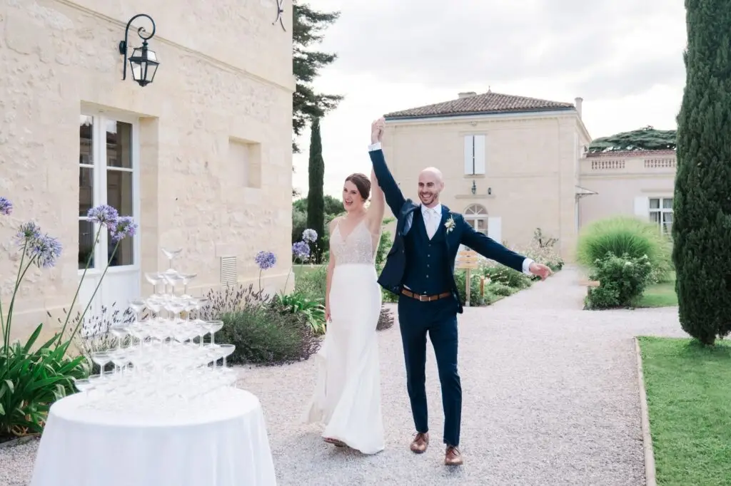 Les mariés font leur entrée main dans la main dans les jardins du Château Gassies à Bordeaux, à côté d’une pyramide de coupes de champagne