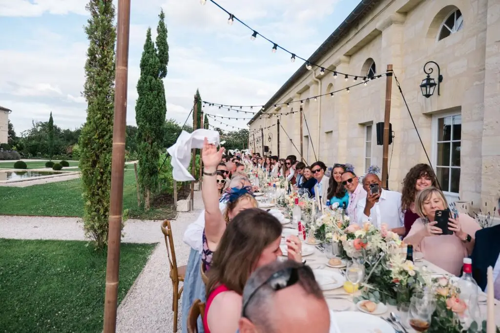 Les invités partagent un dîner de mariage en extérieur autour d’une grande table décorée dans la cour du Château Gassies à Bordeaux, sous des guirlandes lumineuses