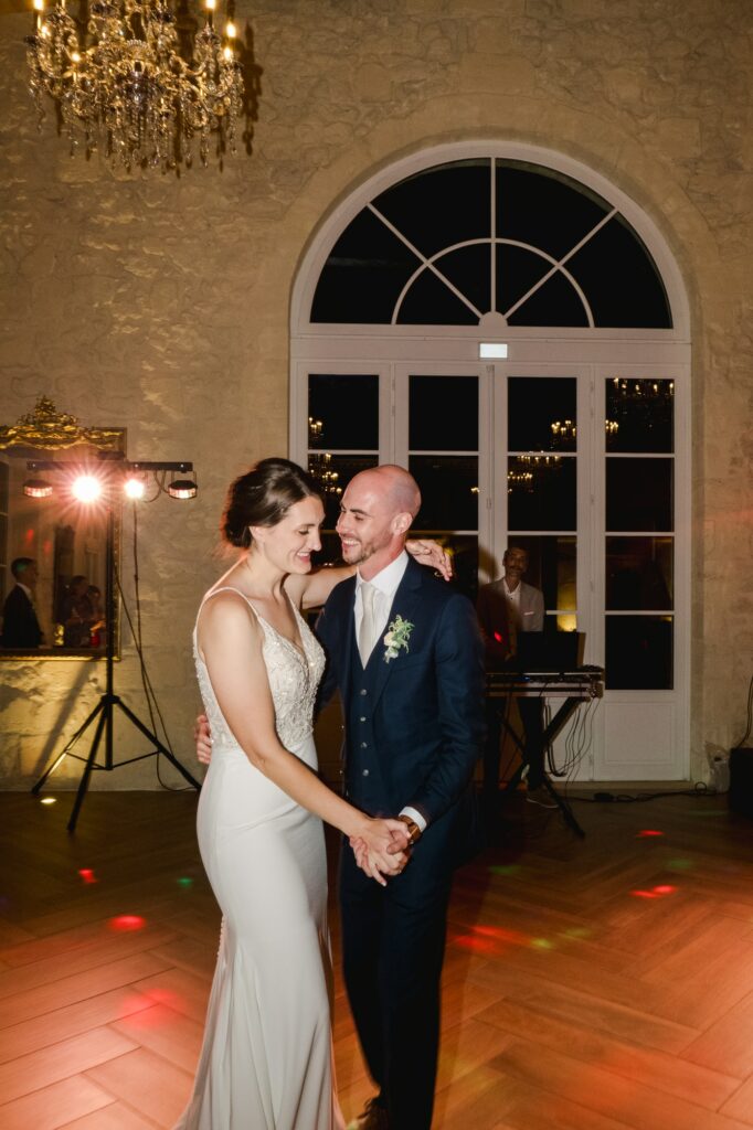 Les mariés se tiennent la main et sourient pendant leur première danse dans une salle de réception éclairée par des lustres.