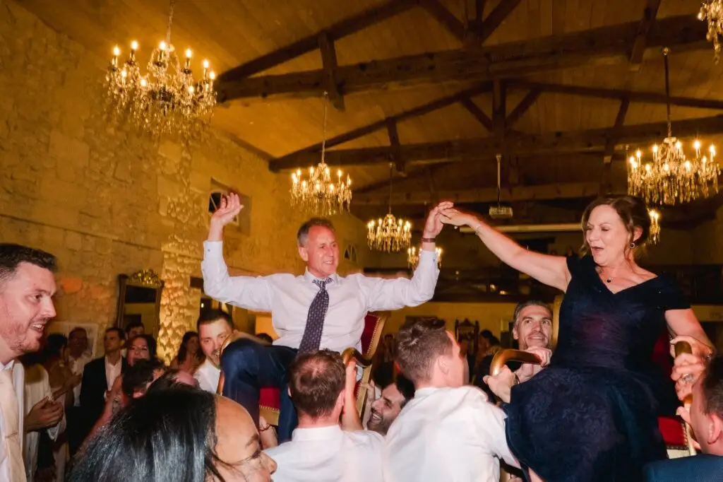 Deux invités sont portés en hauteur sur des chaises par la foule lors d’un moment de célébration pendant la soirée de mariage.