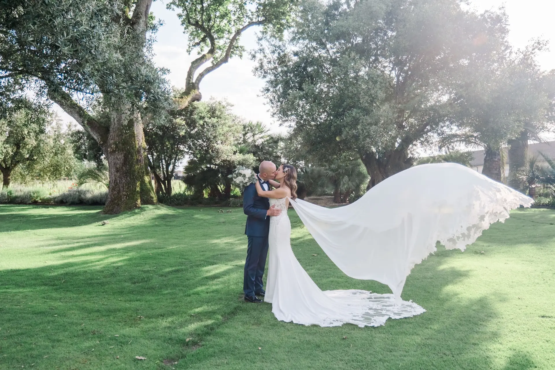 Couple de mariés s’embrassant dans le parc du Château Pape Clément à Pessac, voile de la mariée porté par le vent, photo par Julien Boyer photographe de mariage