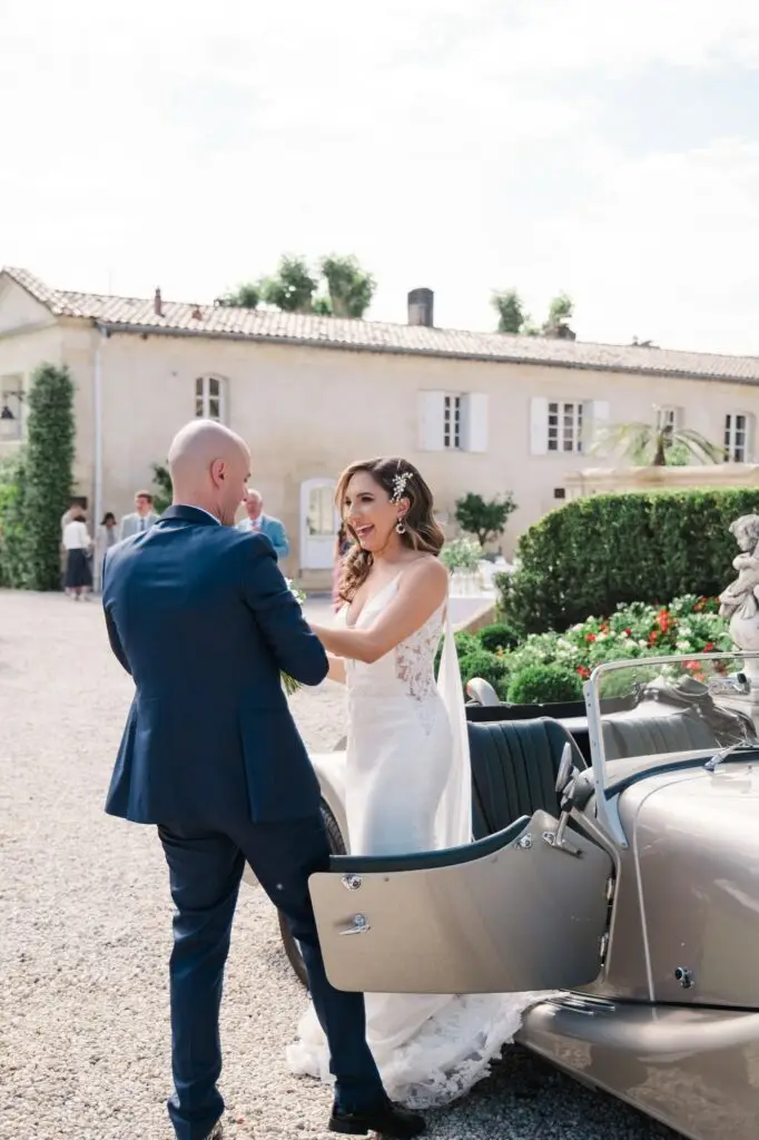 Les mariés descendent d’une voiture de collection dans la cour du Château Pape Clément à Pessac, moment complice photographié par Julien Boyer