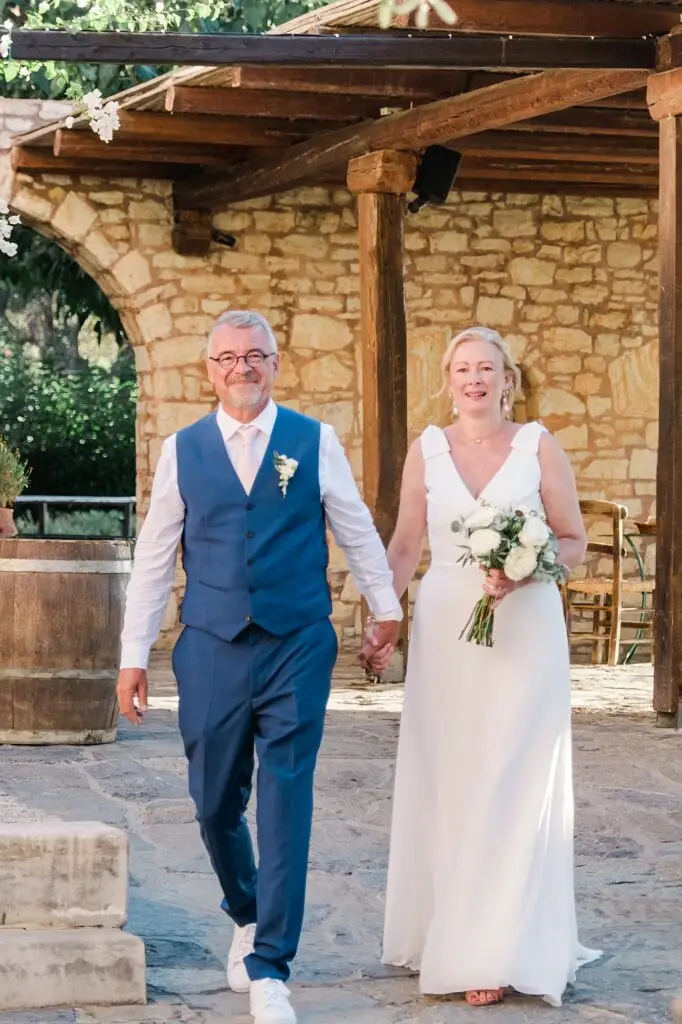 Entrée des mariés main dans la main lors de leur mariage à Agreco Farm près de Rethymnon en Crète, cérémonie en plein air photographiée par Julien Boyer photographe de mariage