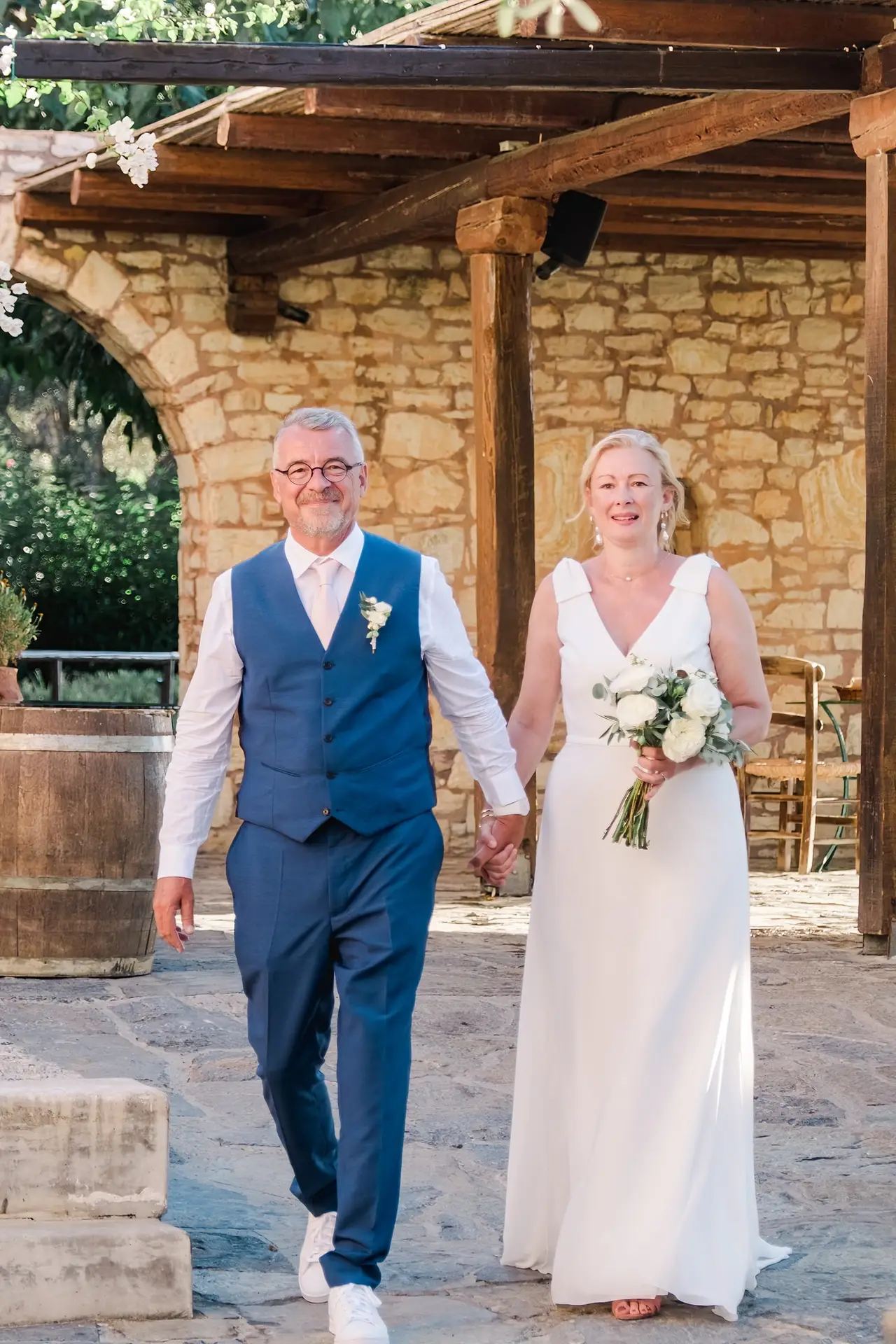 Entrée des mariés main dans la main lors de leur mariage à Agreco Farm près de Rethymnon en Crète, cérémonie en plein air photographiée par Julien Boyer photographe de mariage