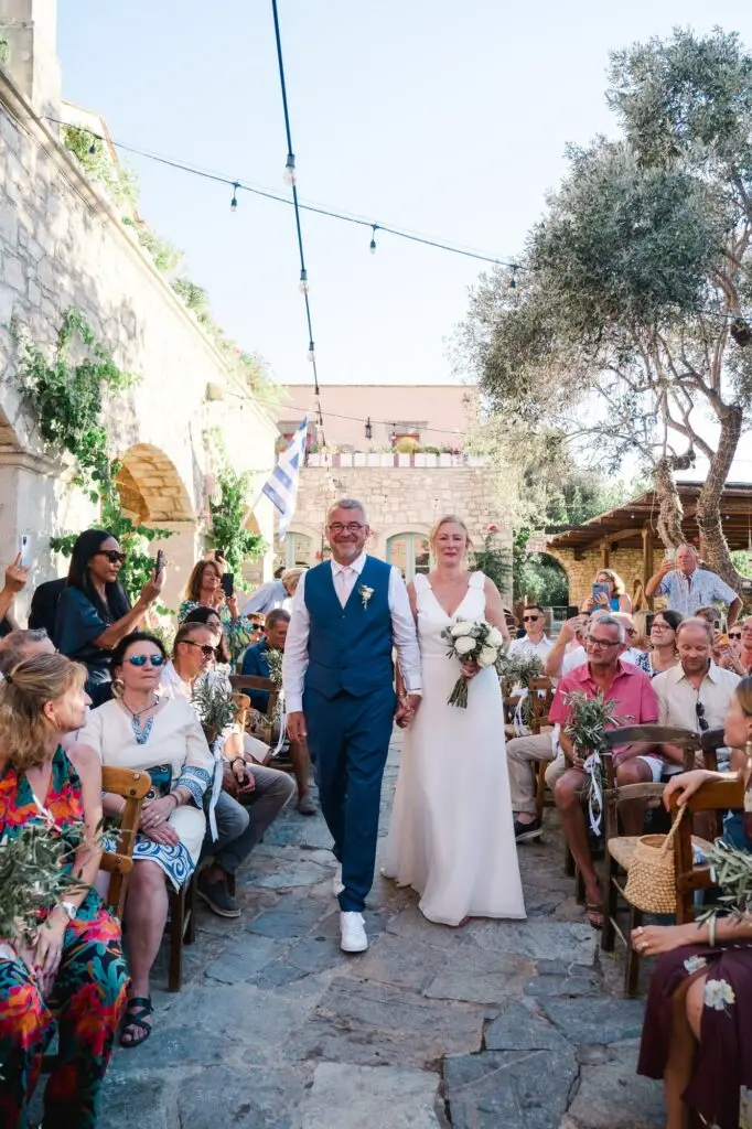 Mariés avançant dans l’allée entourés de leurs invités lors d’une cérémonie de mariage à Agreco Farm en Crète, reportage photo naturel par Julien Boyer