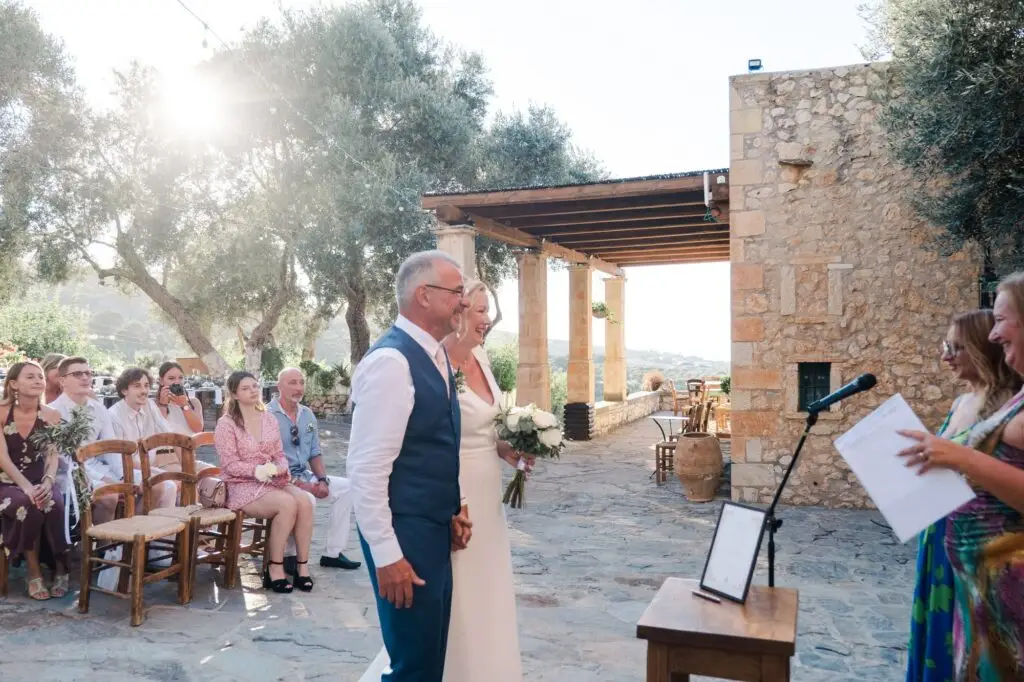 Arrivée des mariés devant l’officiante pour leur cérémonie de mariage à Agreco Farm près de Rethymnon en Crète, lumière naturelle capturée par Julien Boyer photographe de mariage