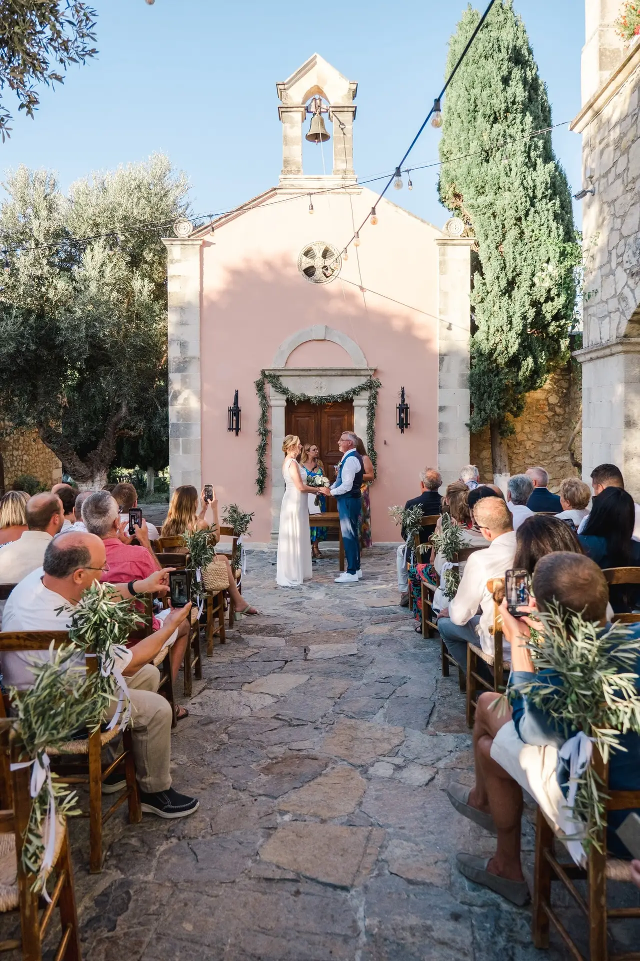 Vue d’ensemble de la cérémonie de mariage à Agreco Farm près de Rethymnon en Crète, mariés devant la chapelle entourés de leurs invités photographiés par Julien Boyer photographe de mariage