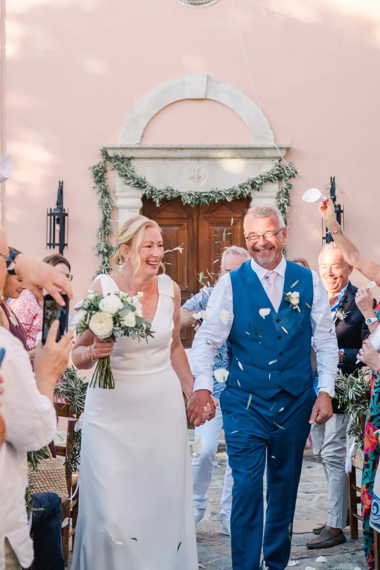 Mariés rayonnants sortant de la chapelle à Agreco Farm en Crète, entourés de leurs invités, photo naturelle par Julien Boyer photographe de mariage