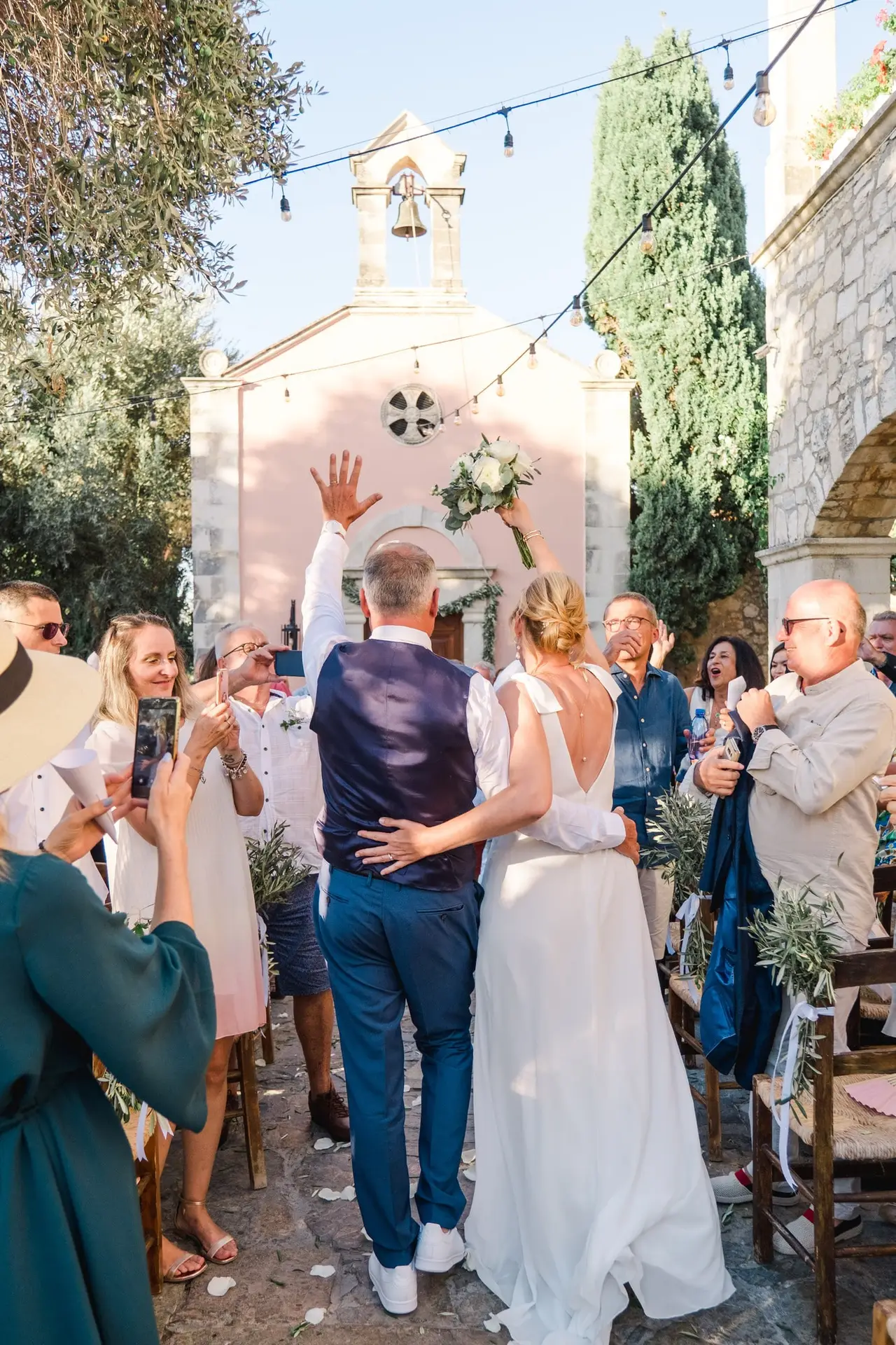 Sortie de cérémonie vue arrière à Agreco Farm en Crète, mariés acclamés par leurs invités devant la chapelle, photo signée Julien Boyer photographe de mariage