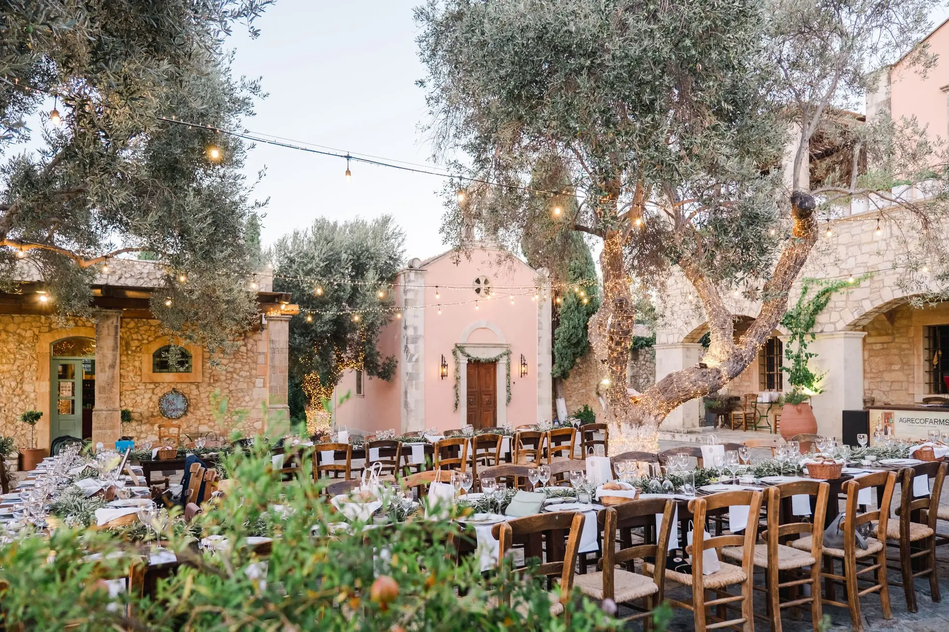 Mise en place de la réception de mariage à Agreco Farm près de Rethymnon en Crète, tables dressées sous les oliviers et guirlandes lumineuses photographiées par Julien Boyer photographe de mariage