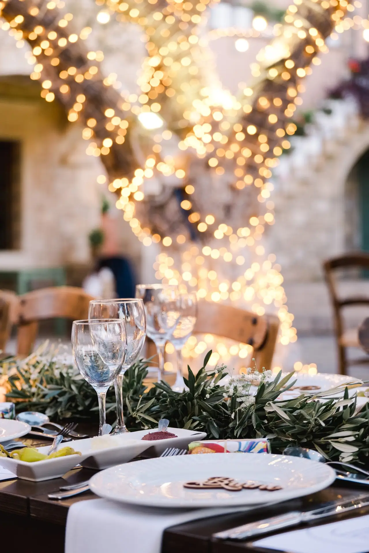 Détail de décoration de table de mariage avec feuillage d’olivier et guirlandes lumineuses autour des arbres, ambiance chaleureuse en Crète photographiée par Julien Boyer