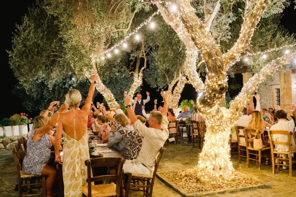 Soirée de mariage festive sous des oliviers décorés de guirlandes lumineuses, invités levant les bras lors d’un mariage en Crète