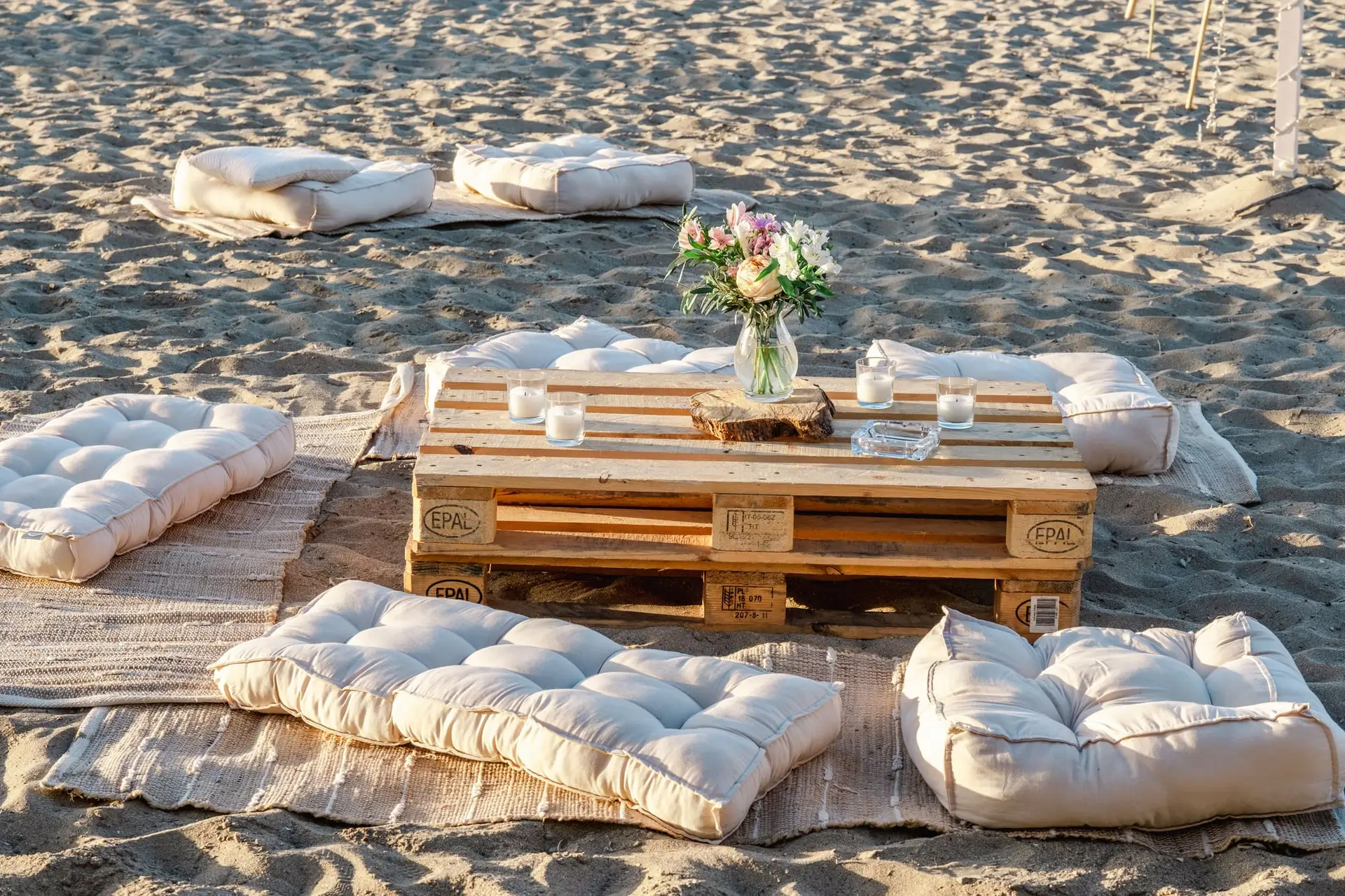 Espace lounge de mariage installé sur la plage avec coussins, palettes en bois et décoration florale pour un mariage en Crète
