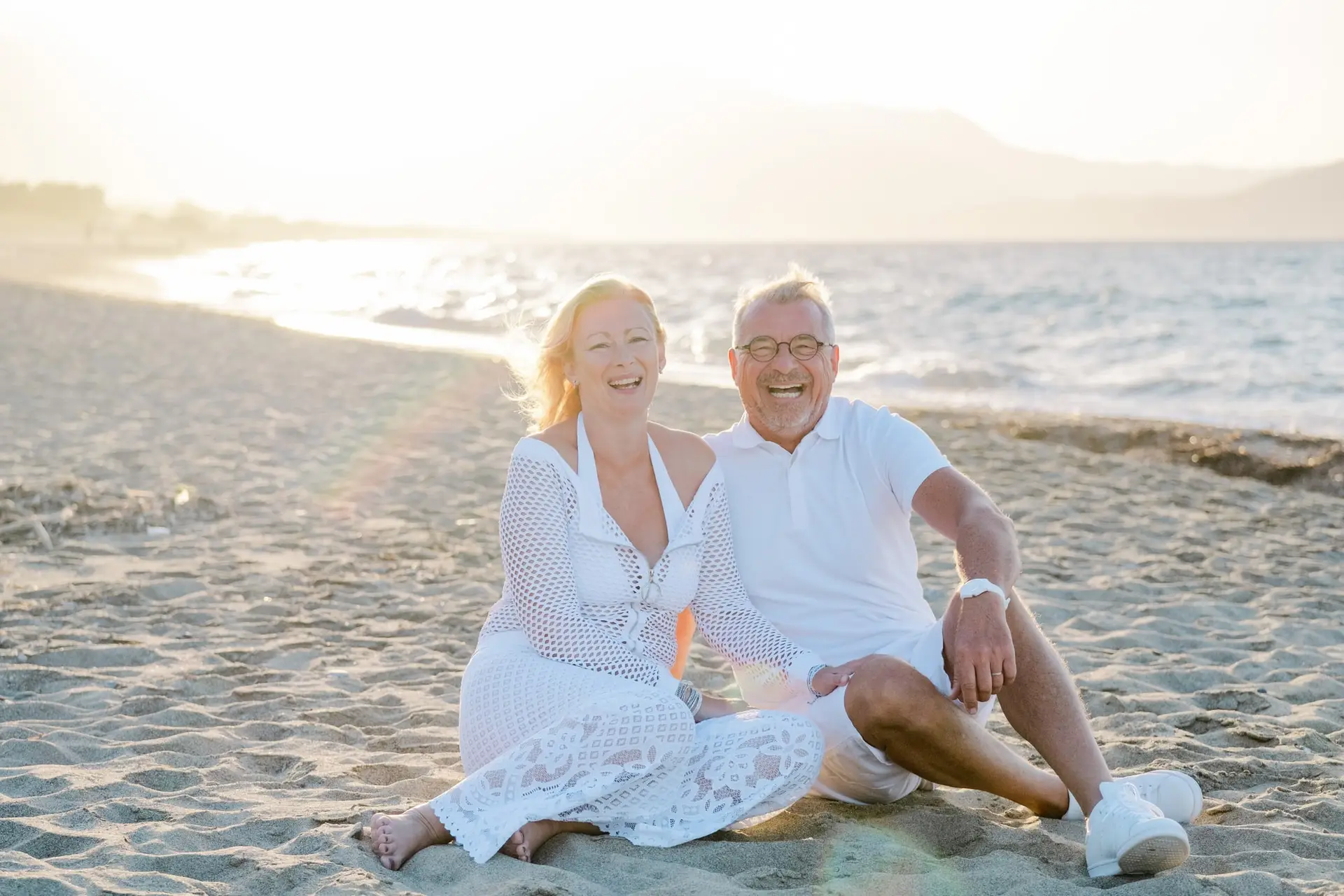 Couple assis sur le sable en bord de mer en Crète, sourires et complicité au coucher du soleil lors d’un mariage destination