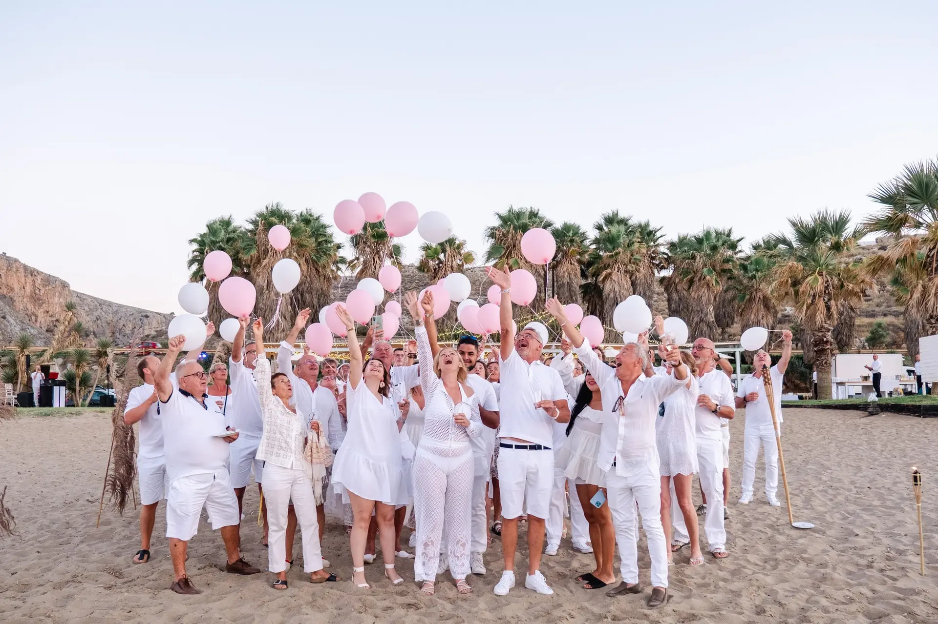 Invités lançant des ballons blancs et roses sur la plage lors d’un mariage en Crète, moment festif et collectif en bord de mer
