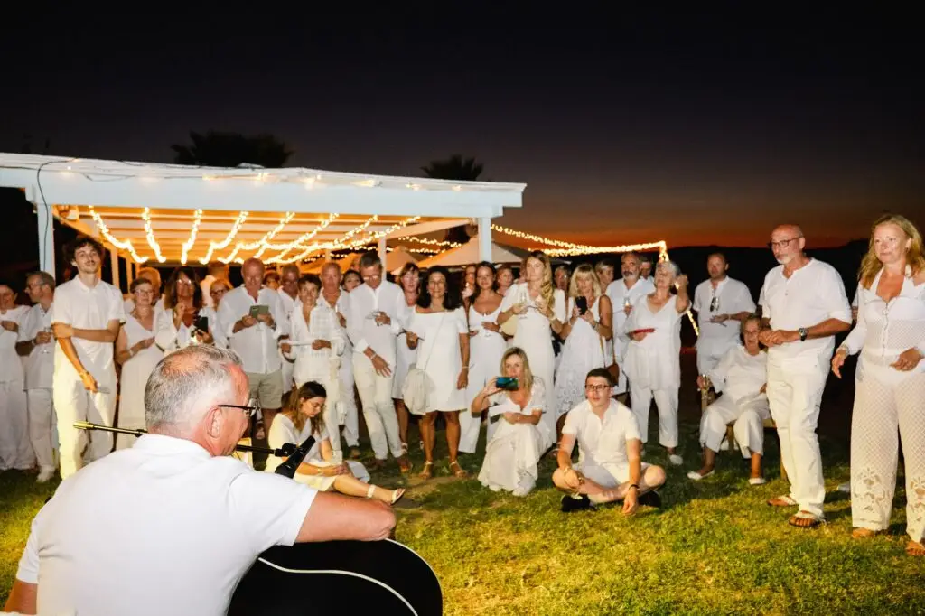 Invités réunis autour d’un musicien pendant la soirée de mariage en extérieur en Crète, ambiance conviviale et festive sous les lumières
