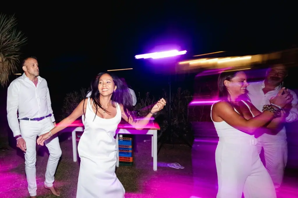 Invités dansant en tenue blanche lors de la soirée de mariage en Crète, avec effets de lumière colorés et ambiance festive en plein air