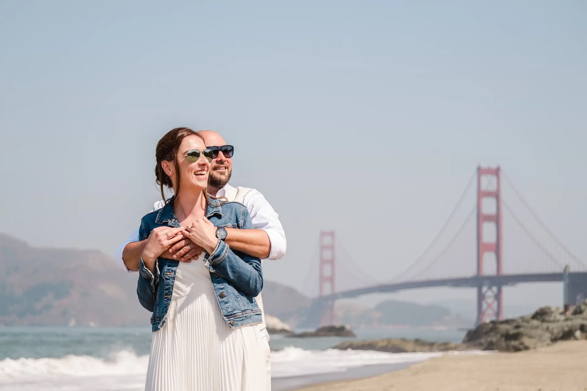 Couple de mariés enlacé et souriant sur la plage à San Francisco avec le Golden Gate Bridge en arrière-plan