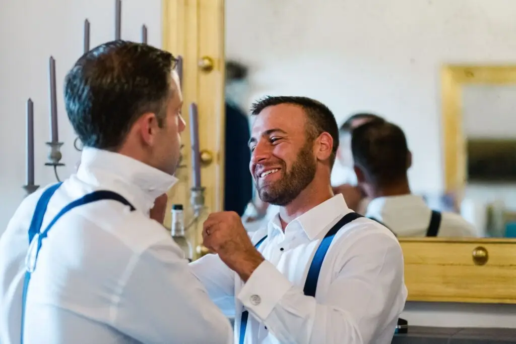 Un témoin aide le marié à ajuster son col avant la cérémonie de mariage à la Villa Vistarenni en Toscane, photographie de mariage haut de gamme.