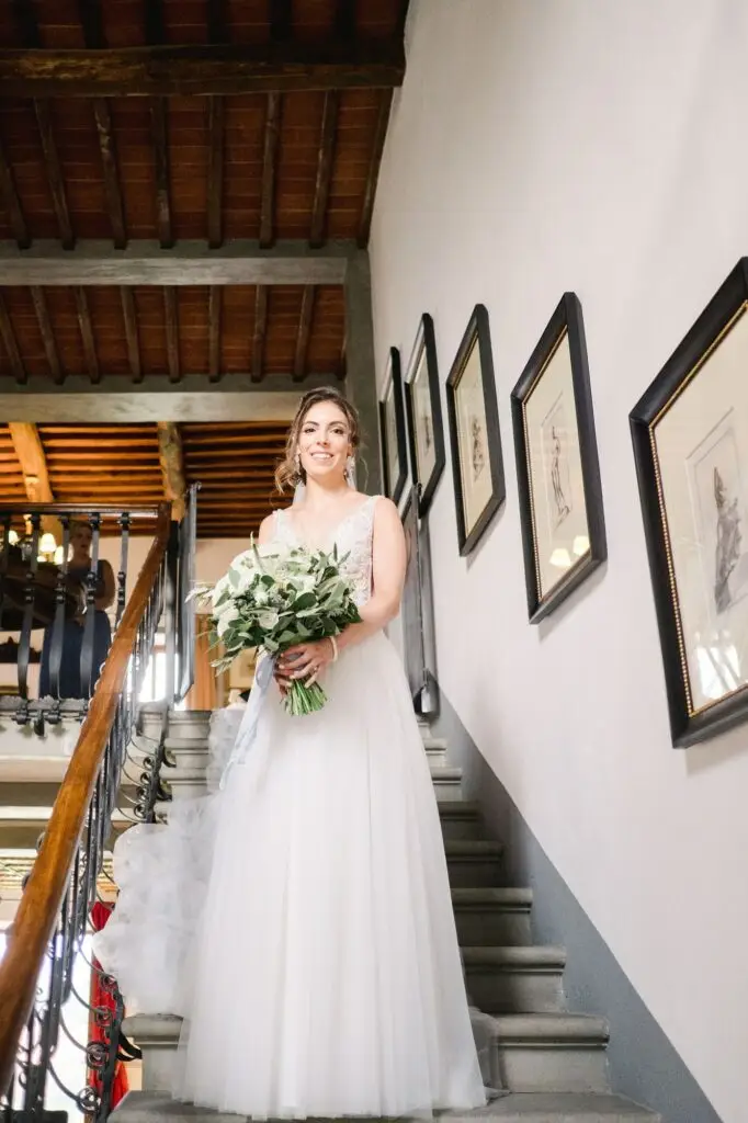 Mariée descendant un escalier intérieur avec son bouquet lors d’un mariage à la Villa Vistarenni en Toscane, scène élégante et lumineuse.