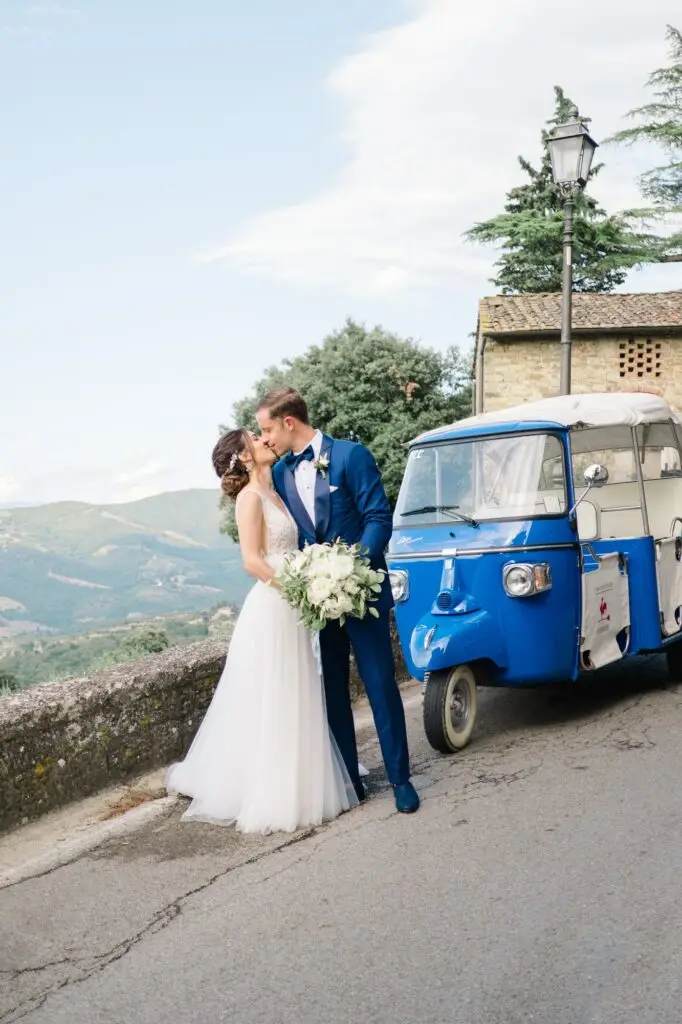 Les mariés s’embrassent devant un Piaggio bleu avec vue sur la Toscane, séance photo de mariage élégante en Italie, photo prise par Julien Boyer.
