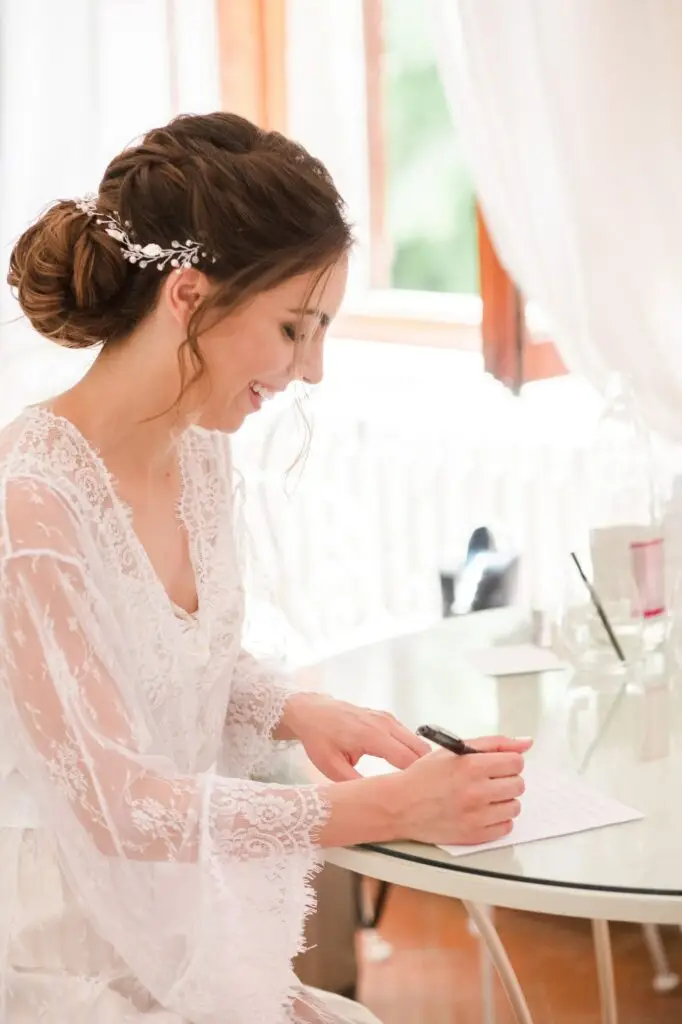 Mariée en robe de dentelle écrivant une lettre pendant ses préparatifs à la Villa Vistarenni en Toscane, scène lumineuse et pleine d’émotion.